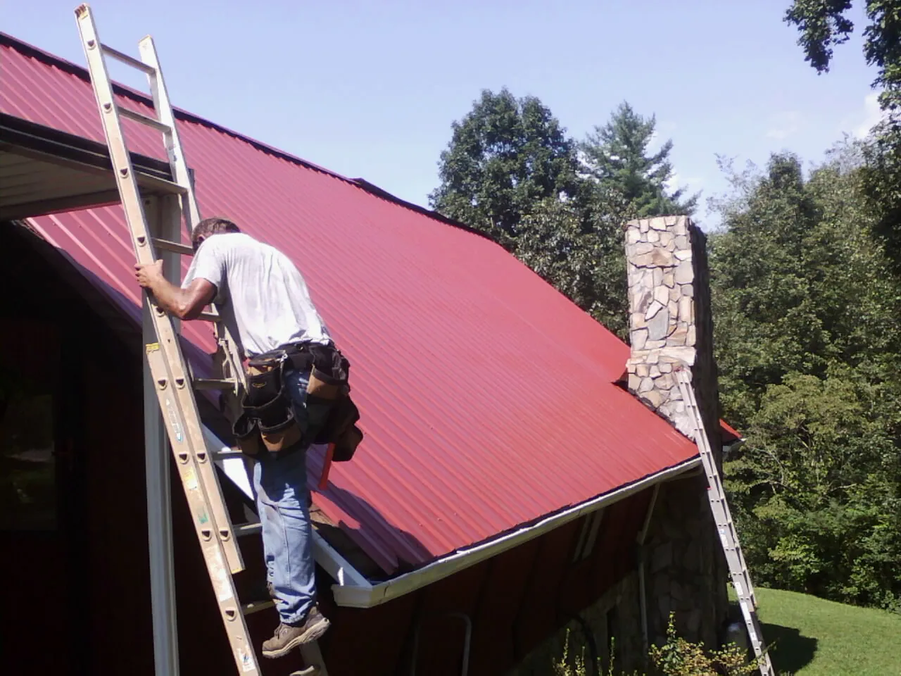 Roofer on a ladder beside a red metal roof and stone chimney — Soffit & Fascia Repair by Roofing Brothers in Waretown